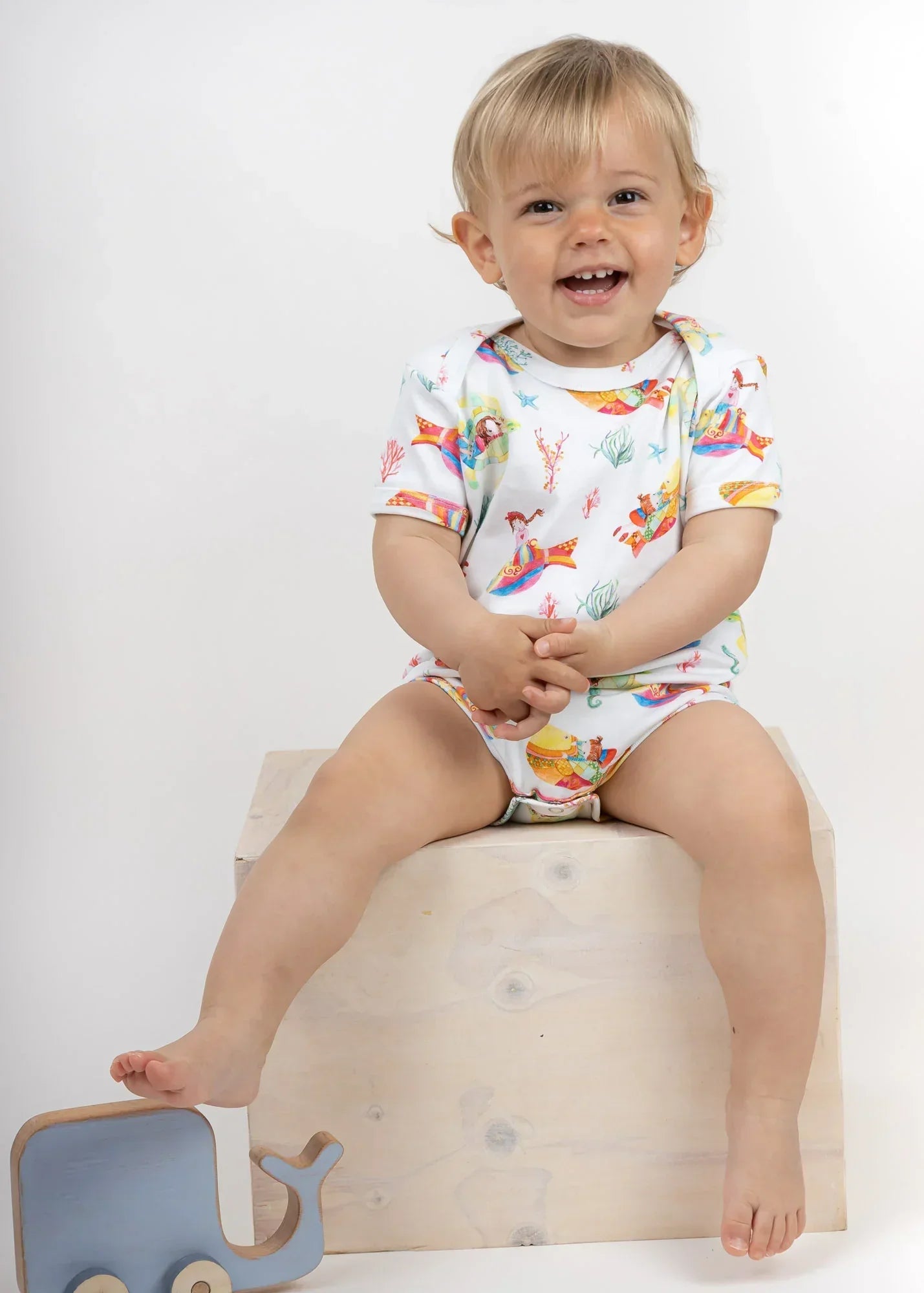 Smiling toddler wearing a colorful ZOE baby bodysuit with playful print, seated on a wooden box