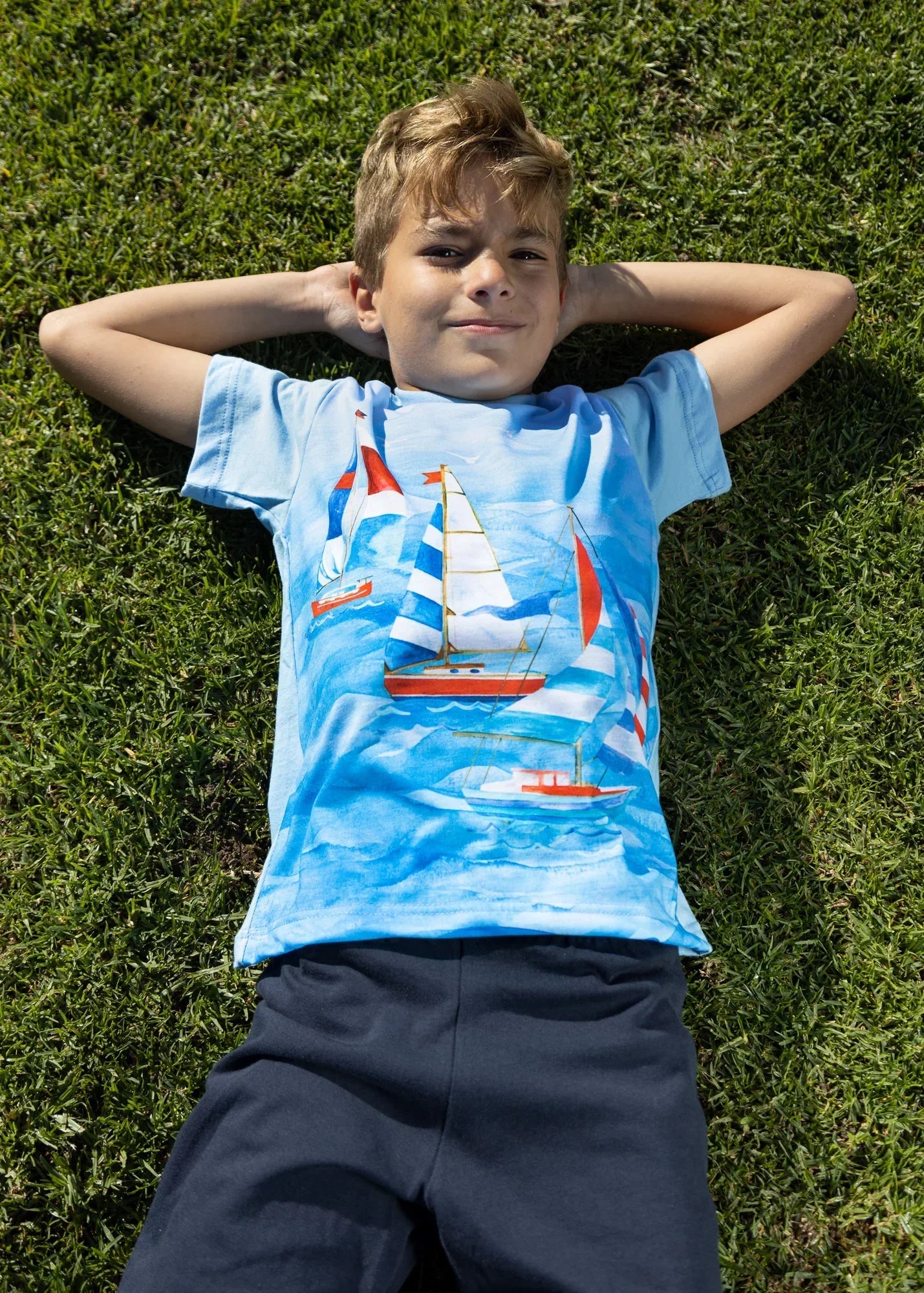 Boy wearing ZOE Greek cotton sailboat T-shirt lying on grass, outdoor summer fashion