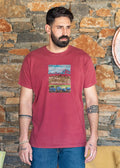 Man wearing ZOE maroon T-shirt with original art, standing by rustic stone wall, Greek casual fashion
